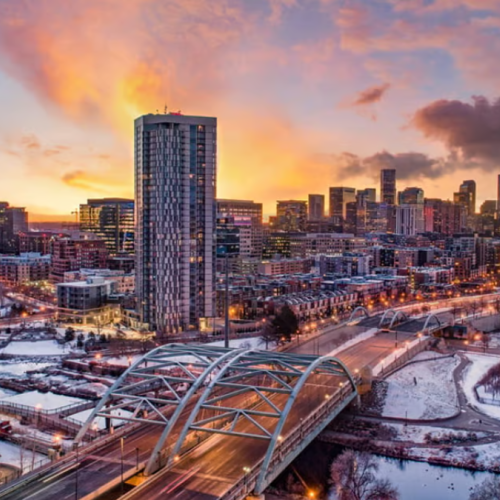 Image of a sunset, with a skyscraper and a bridge with snow on the ground. 