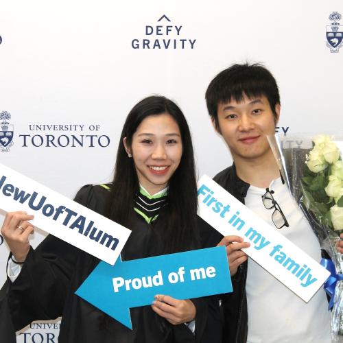 A new grad celebrated graduation at U of T with their friend, holding signs and flowers in front of a “Defy Gravity” backdrop.