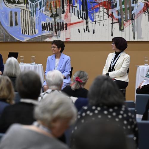 "Four women sit at the front of a room engaged in a panel discussion at St. Michael’s College, University of Toronto. One of the women speaks into a microphone while the others listen attentively. They are seated in front of a colorful abstract painting, with round tables holding water glasses beside them. An audience of older adults listens from rows of chairs.