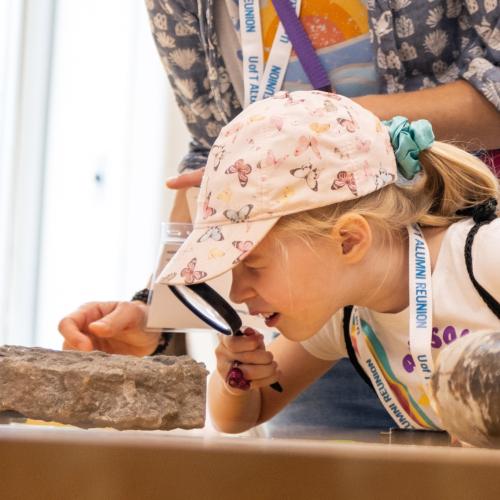 A young child, accompanied by an adult, examines a rock through a magnifying glass.