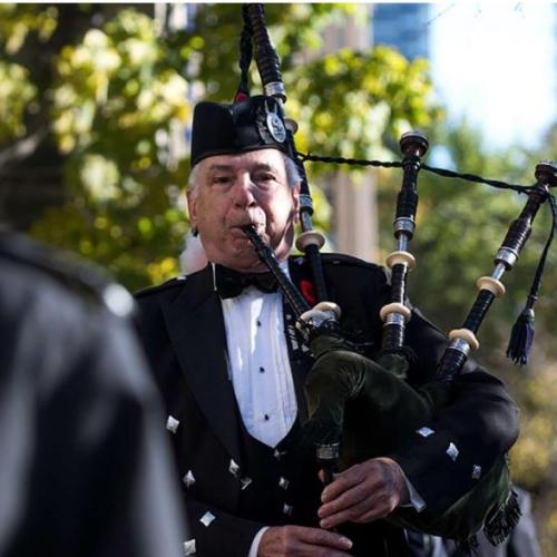 Man playing bagpipe, with trees behind him. Man dressed in black with white shirt. 