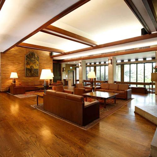Image of Massey College's Common Room. There is brown furniture placed in the room to invite conversation. There are tables, lamps and a fireplace with large windows. Brick walls and tall white ceiling.