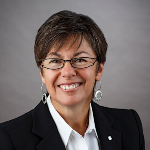 Photo of speaker Kim Pate wearing black jacket, white shirt, glasses against a grey background. 