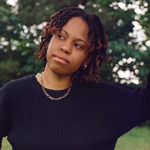 Isabel Okoro standing outside leaning their arm on a tree, while looking at the view of nature. 