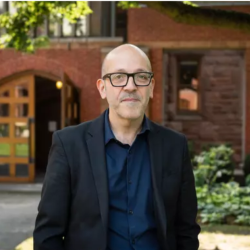 Image of man in shirt, suit and black rimmed eye glasses, standing outside with trees and building in background. 