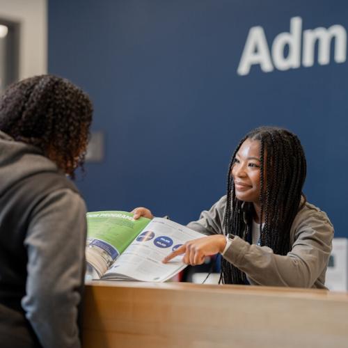 Two people talking, with one person with a booklet behind a desk showing directions to the person across them. 