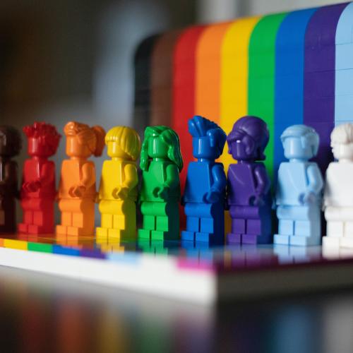 Rainbow colored Lego characters standing in front of a Pride flag background